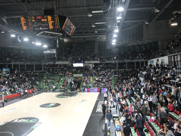 Große Halle mit Zuschauern bei einem Basketballspiel, mit sichtbarem Basketballkorb, Sitzbereichen und Überwachungsbildschirmen und Beleuchtung an der Decke.