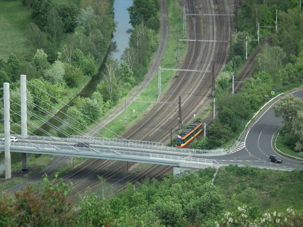 Ein Zug überquert eine Brücke über einen Fluss, mit Fahrzeugen auf der Straße darunter und Stromleitungen, die entlang der Brücke verlaufen, vor einem Hintergrund aus Bäumen und Gras.