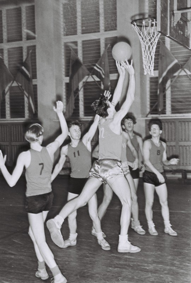 Schwarzes Foto von jungen Männern, die Basketball in einer Turnhalle spielen, mit einem Basketballkorb in der rechten oberen Ecke und Flaggen im Hintergrund.