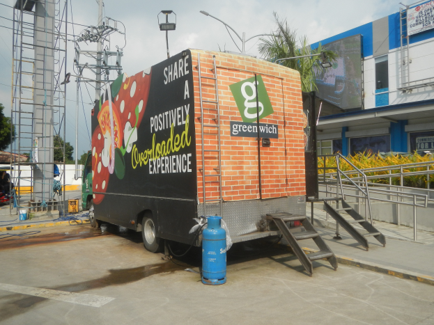 Ein Lastwagen parkt vor einem Gebäude mit einer Tafel, auf der "Teilen Sie eine positiv überladene Erfahrung" steht, mit Stufen, die hinaufführen, einem Zylinder auf dem Boden, Geländern, Strommasten mit Drähten, Laternen, Bäumen und einem bewölkten Himmel im Hintergrund.