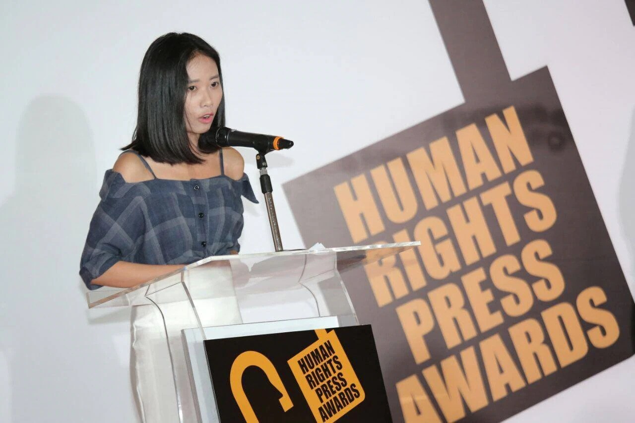 Eine Frau in einem schwarzen Kleid mit einem Dutt steht an einem Rednerpult mit einem Mikrofon und spricht bei der 'Human Rights Press Awards'-Veranstaltung, ihr Gesichtsausdruck ist ernst und konzentriert.