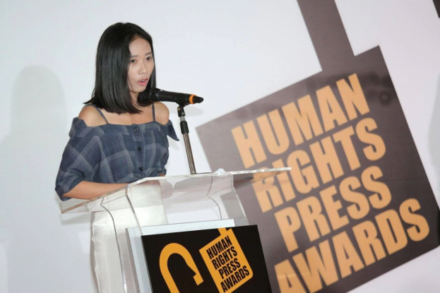 Eine Frau in einem schwarzen Kleid mit einem Dutt steht an einem Rednerpult mit einem Mikrofon und spricht bei der 'Human Rights Press Awards'-Veranstaltung, ihr Gesichtsausdruck ist ernst und konzentriert.