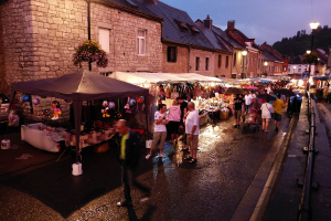 Eine Gruppe von Menschen, die auf einer Straße mit Geschäften, Schirmen, hängenden Spielzeugen, Taschen, Gebäuden mit Fenstern, Bäumen, Straßenlaternen und Barrieren geht.