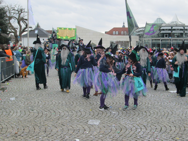 Eine Gruppe von Menschen in Hexenkostümen, die schwarze und lila Kleider und Hüte tragen und einige Fahnen halten, gehen eine Kopfsteinpflasterstraße entlang, neben einem Geländer und Gebäuden unter einem klaren blauen Himmel.
