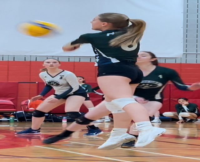 Eine Gruppe junger Frauen beim Volleyballspielen auf einem Platz mit einem Ball in der Luft, einer Wand im Hintergrund und verschiedenen Gegenständen wie Stühlen, Taschen, Flaschen und Menschen in Turnschuhen.