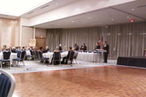 Eine Gruppe von Menschen, die auf Stühlen sitzen und stehen, mit Flaggen, Lichtern, Schildern und Vorhängen im Hintergrund.