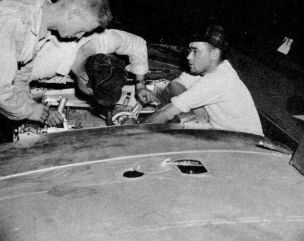 Drei Männer in Mützen reparieren ein schwarz-weißes Auto in einer Garage umgeben von Werkzeugen und Materialien.