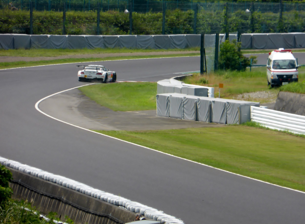 Ein Rennwagen, der auf einer Rennstrecke fährt, mit einem Polizeiauto im Hintergrund, umgeben von Gras, Pflanzen, Bäumen und Absperrungen, mit einem Fahrzeug auf der rechten Seite und Text unten.
