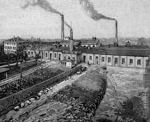 Schwarze und weiße Zeichnung einer Fabrik mit Rauch aus Schornsteinen, umgeben von Gebäuden, Bäumen, Pflanzen, Pfählen und Bahnschienen, beschriftet mit "1892 - die Fabrik des Eisenerzes" unten.