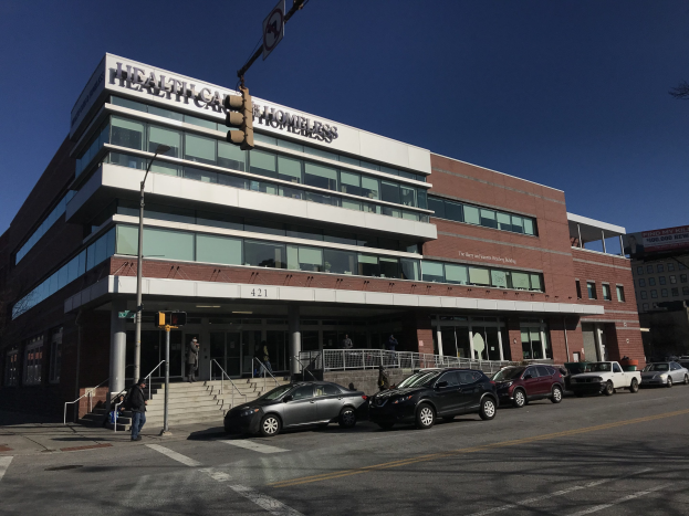 Großes Gebäude mit der Aufschrift 'New York City Health Care Homeless Center' mit parkenden Autos, Passanten, Straßenmöbeln und Bäumen unter einem klaren Himmel.