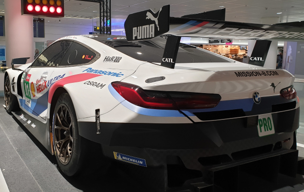 BMW M8 GT3 Rennauto in einer Ausstellungshalle mit einem Rad im Vordergrund.