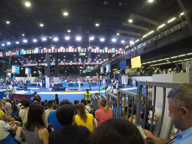 Große Menschenmenge sitzt und steht in einer Indoor-Sportarena mit Bildschirmen, Fahnen und Deckenbeleuchtung im Hintergrund.