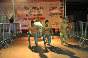 Drei Jungen ohne Hemden knien während eines kulturellen Ereignisses auf dem Boden, mit Grills auf beiden Seiten und einem großen Banner im Hintergrund, während eine Person auf der linken Seite ein Foto mit einer Kamera macht, unter Deckenbeleuchtung.