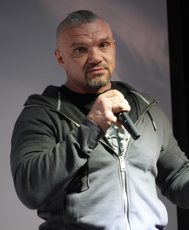 Ein Mann in einer schwarzen Kapuzenjacke und einem schwarzen Anzug mit einem weißen Hemd, der ein Mikrofon hält, steht selbstbewusst vor einer Wand und sieht aus wie ein Comedian mit zurückgekämmtem Haar.