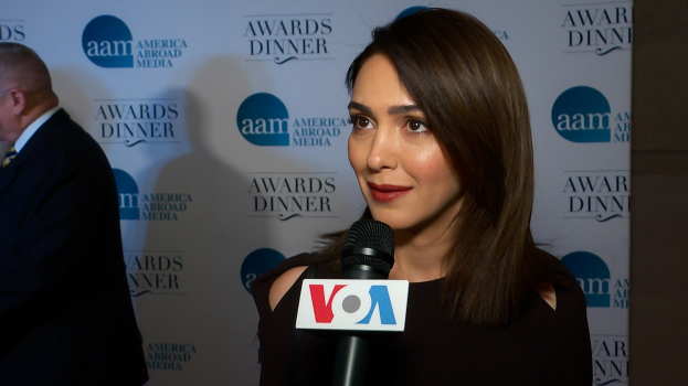 Emmy Rossum in einem schwarzen Kleid hält ein Mikrofon, neben einem Mann mit einer Tafel 'Aam Awards Dinner' im Hintergrund.