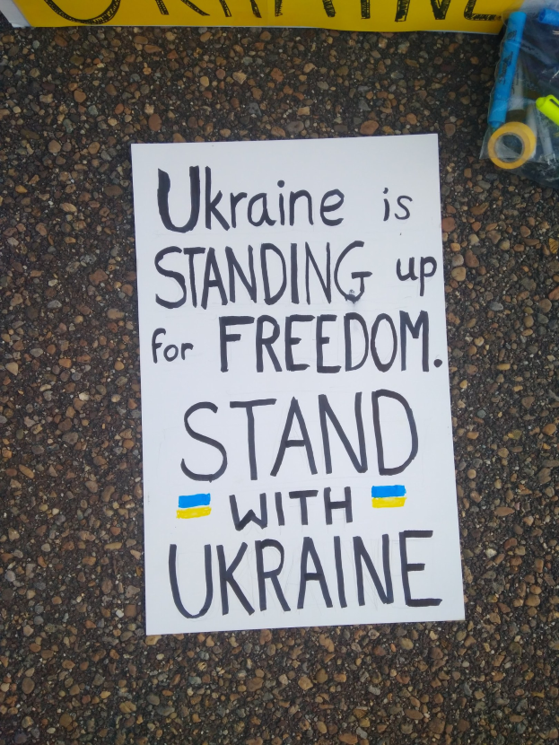 Weißees Papier mit "Ukraine steht für Freiheit, steht mit Ukraine" Text auf dem Boden vor einem gelben Banner, mit einer Plastikabdeckung mit verschiedenen Gegenständen rechts.