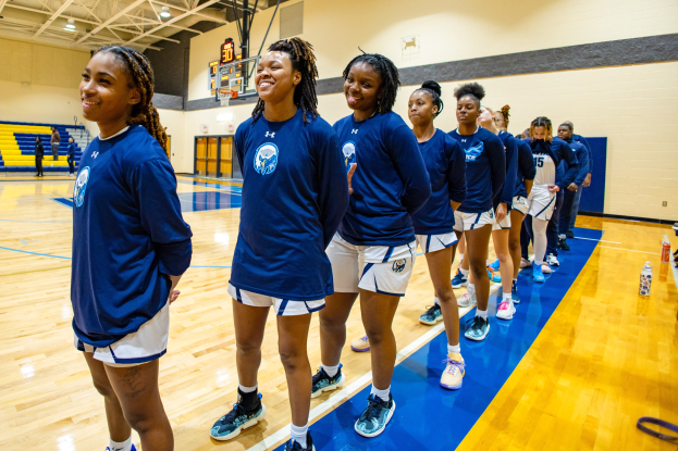 Eine Gruppe junger Frauen in blauen T-Shirts, die auf einem Basketballfeld lächeln, mit Flaschen und Gegenständen auf dem Boden, Menschen im Hintergrund und einem Basketballkorb
