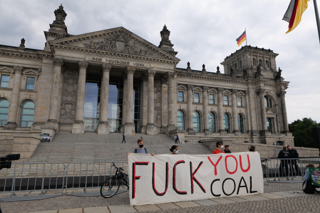 Menschen mit einem "Fuck You Coal"-Schild vor dem Reichstaggebäude in Berlin, mit den architektonischen Details des Gebäudes, einem Fahrrad, Bäumen, einer Flagge und einem bewölkten Himmel.