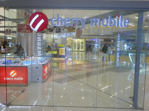 Cherry Mobile Store in einem Einkaufszentrum mit Glaswänden und -türen, Menschen, die gehen, Deckenleuchten und Tische mit Gegenständen im Hintergrund.