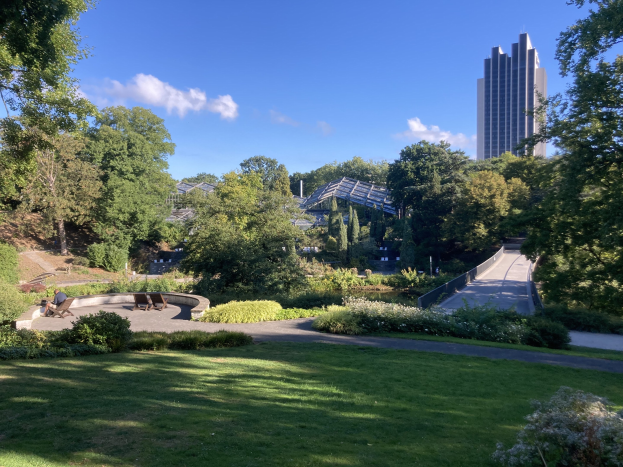 Ein Park mit grünem Gras, Bäumen, Pflanzen, Blumen, Bänken und einem Weg, vor einer Kulisse aus Gebäuden und einem bewölkten Himmel.