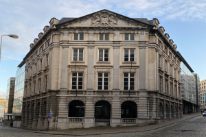 Das Bankgebäude in Frankfurt, ein großes Gebäude mit vielen Fenstern, umgeben von Straßenlaternen, Autos und Passanten, mit einem bewölkten Himmel im Hintergrund.