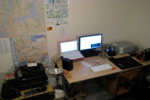 Ein Raum mit einem Tisch, der einen Laptop, einen Lautsprecher, einen Monitor, eine Tastatur, einen Ball, eine Maus, einen Scanner und Papiere enthält; ein weiterer Tisch mit Elektronikgeräten ist links unten sichtbar, mit Charts und Postern an der Wand dahinter.