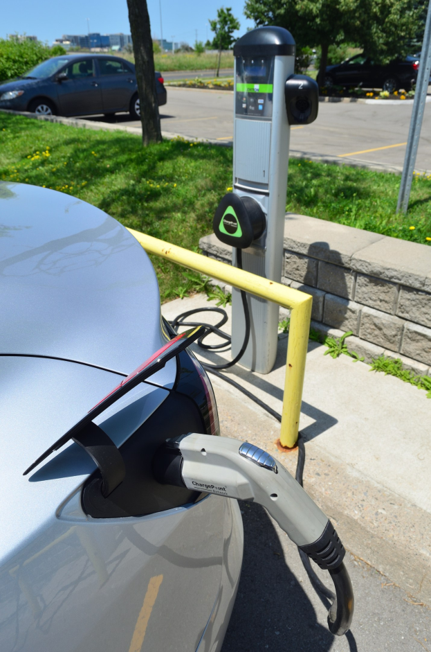 Ladestation für Elektroauto am Straßenrand mit Gras, Bäumen, Strommasten und Gebäuden im Hintergrund unter einem klaren blauen Himmel.