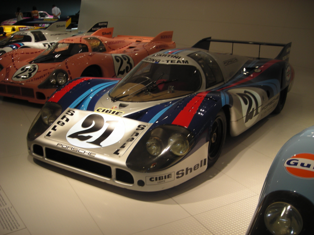 Ein Porsche 917K Le Mans Rennwagen in einem Museum ausgestellt, umgeben von anderen Sportwagen und Menschen, mit einer Wand und informativem Text im Hintergrund.