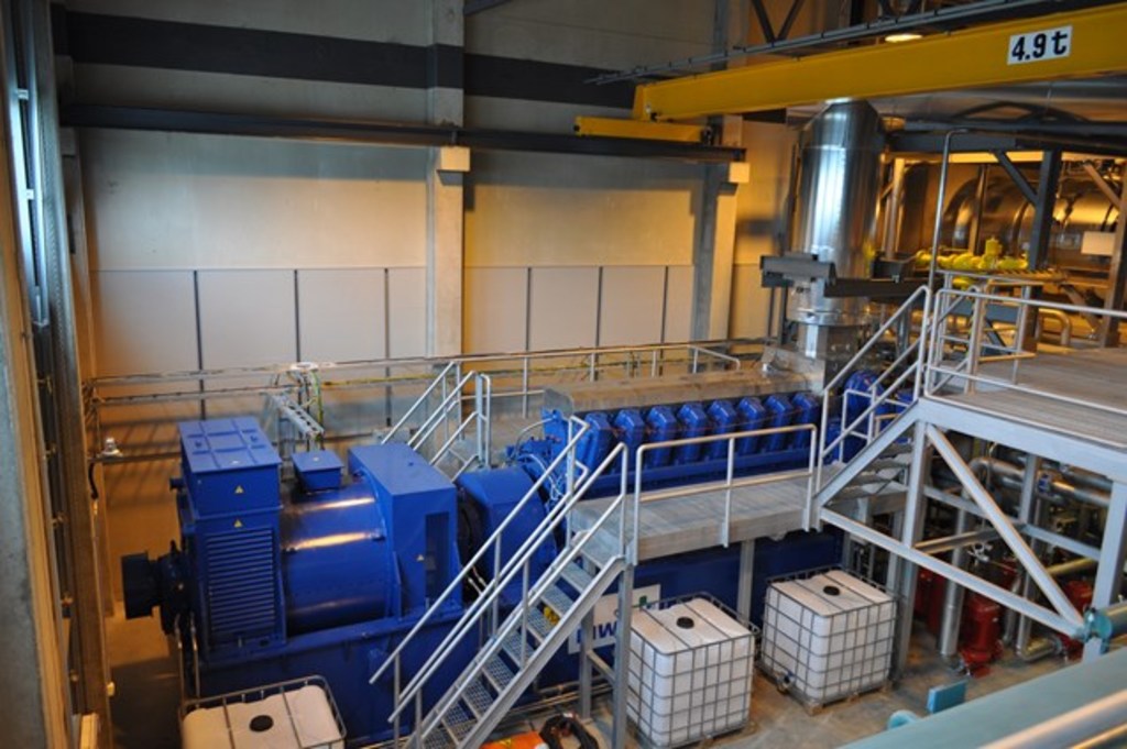 Ein Fabrikinneres mit Trägern, einer Treppe mit Geländer, Containern, Lampen, Rohren und Wänden.