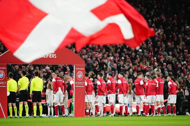 Menschen auf einem Fußballfeld mit einer roten und weißen Fahne im Vordergrund, ein Bogen mit der Aufschrift "Bayern München vs. Bayern München Wetten & Vorschau" im Hintergrund und eine große Menschenmenge im Stadion.