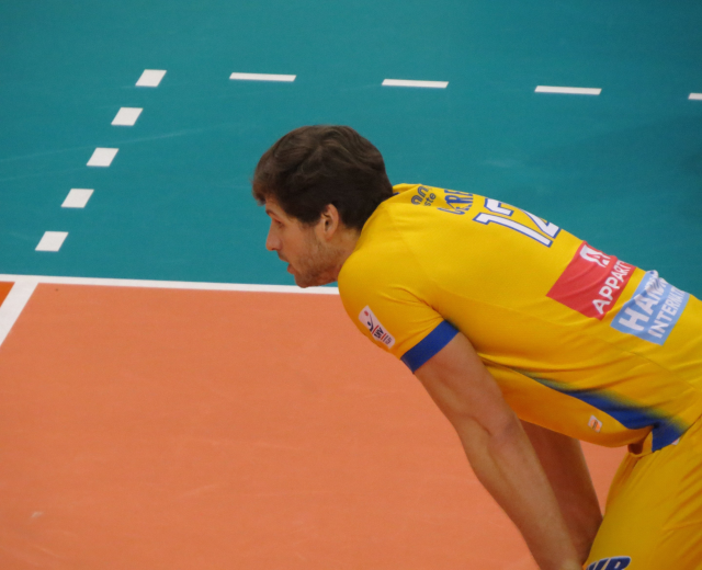 Eine professionelle Volleyballspielerin in einem gelben "Volleyball"-T-Shirt steht selbstbewusst auf einem Court und bereitet sich darauf vor, den Ball zu servieren.