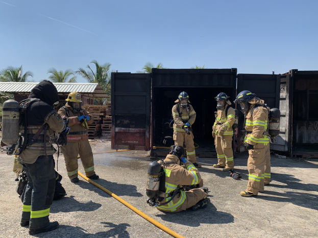 Feuerwehrleute in Helmen und Ausrüstung vor einem Gebäude mit Rohren stehend, mit Bäumen, Schuppen und hölzernen Gegenständen im Hintergrund während einer scheinbaren Brandbekämpfungstrainingssitzung.