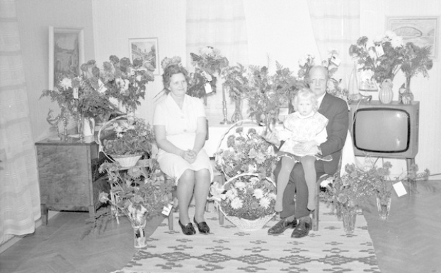 Schwarzes und weißes Foto einer Familie aus drei Personen - einem Mann, einer Frau und einem Kind - die auf einem Sofa sitzen, vor einem Fernseher. Blumenvasen stehen auf Tischen, ein Teppich bedeckt den Boden und Vorhänge und Fotoalben sind im Hintergrund zu sehen.