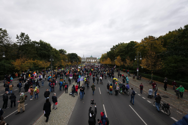 Eine große Gruppe von Menschen marschiert eine baumbestandene Straße in Berlin entlang, mit Kameras in der Hand, vor einem Gebäude und einem klaren Himmel.