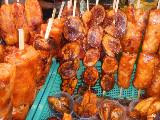 Marktstand mit Spießen von Hähnchenflügeln, verschiedenen Lebensmitteln in Plastikbehältern und Menschen im Hintergrund.