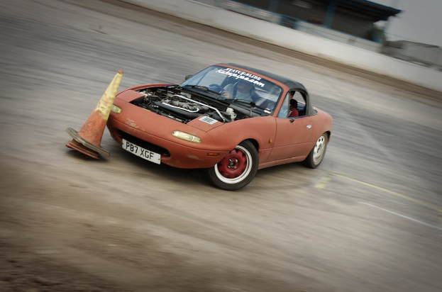 Eine Person, die ein rotes Auto auf einer Rennstrecke fährt, mit einem Verkehrskegel vorneweg, der Hintergrund durch hohe Geschwindigkeit unscharf.