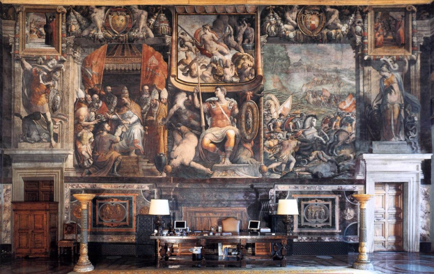 Ein großer Raum mit einem prominenten Gemälde an der Wand, ein Tisch mit Gegenständen, zwei Lampen zu beiden Seiten, Türen links und rechts und ein Teppichboden, befindet sich im Palazzo della Signoria in Florenz, Italien.