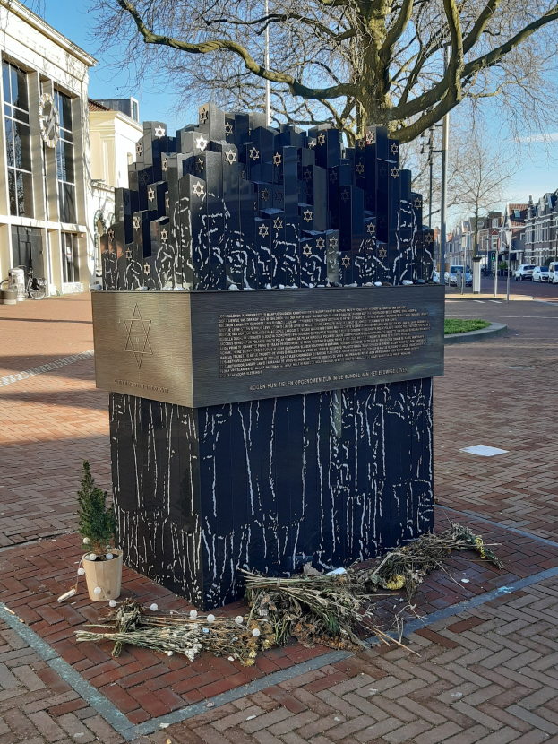 Denkmal in einem Stadtplatz gewidmet an Holocaust-Opfer, mit einem Baum davor, einer Topfpflanze, verstreuten trockenen Blättern und Gebäuden, Fahrzeugen, Polen und Bäumen im Hintergrund.