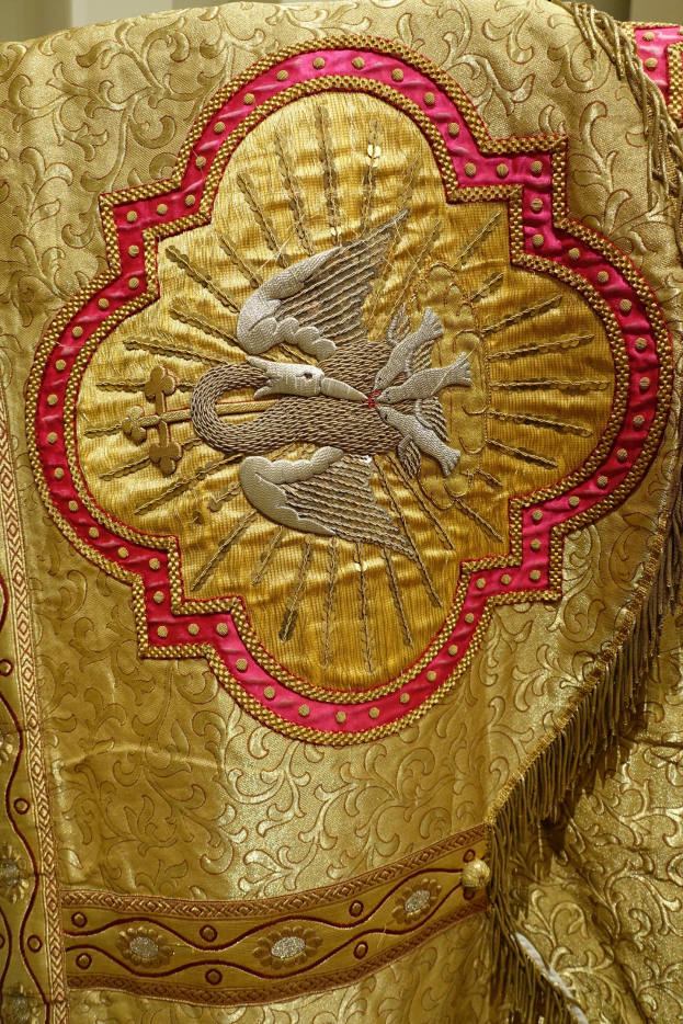 Nahaufnahme eines Priestergewandes mit aufwendiger Gold- und Rotstickerei auf einem reichhaltigen Stoff, vor einem neutralen Wandhintergrund.