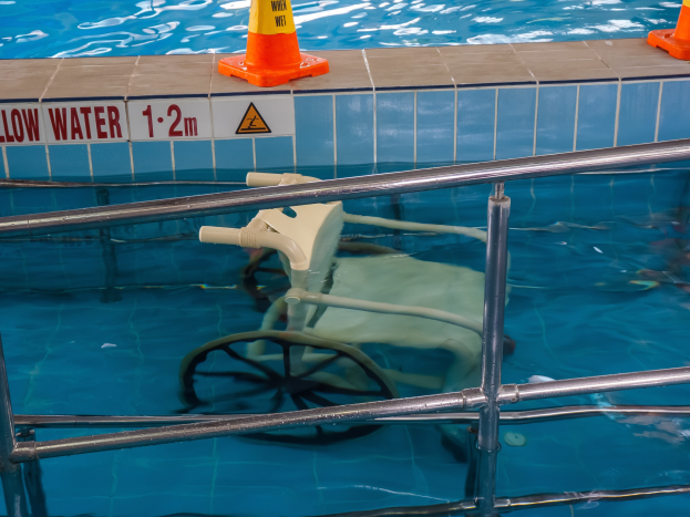 Schwimmbad mit einem Stuhl in der Mitte, umgeben von Sicherheitstonnen und einem Geländer, sowie einem Schild an der Wand, das verfügbare Schwimmausrüstung anzeigt.