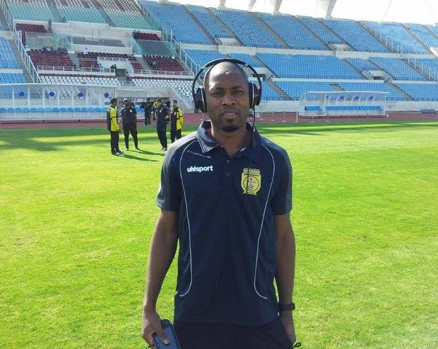 Ein Mann in einem Headset, wahrscheinlich der Trainer der ghanaischen Nationalmannschaft, steht auf einem Fussballfeld in der Nähe eines Stadions und hält ein Objekt, während im Hintergrund Menschen zu sehen sind und der Himmel sichtbar ist.