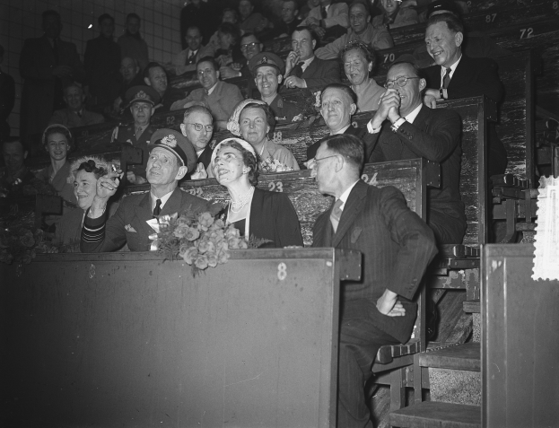 Schwarzes und weißes Foto einer Menschenmenge in einem Stadion mit Sitzenden und Stehenden, einem Podium mit Blumenstrauß, Stufen auf der rechten Seite und einer Wand im Hintergrund.