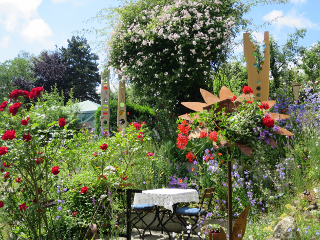 Ein Garten auf der Chelsea Flower Show mit bunten Blumen, Pflanzen, einem Tisch mit einer Decke, Stühlen, Bäumen und einem bewölkten Himmel.