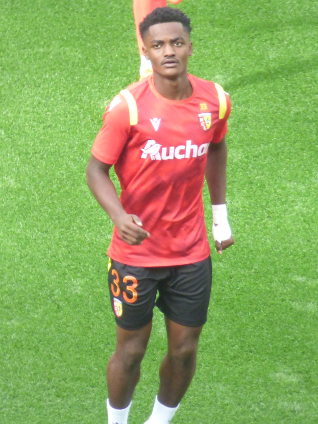 Emmanuel Adebayor, wearing a red shirt and black shorts, walks on a soccer field with two people standing in the background.