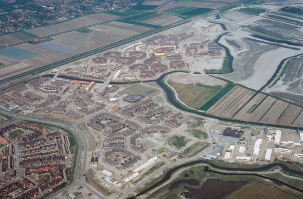 Luftaufnahme einer Stadt mit dichtem Gebäudebestand, Straßen, Feldern, Bäumen und einem zentralen Reservoir umgeben von urbanen und grünen Bereichen.