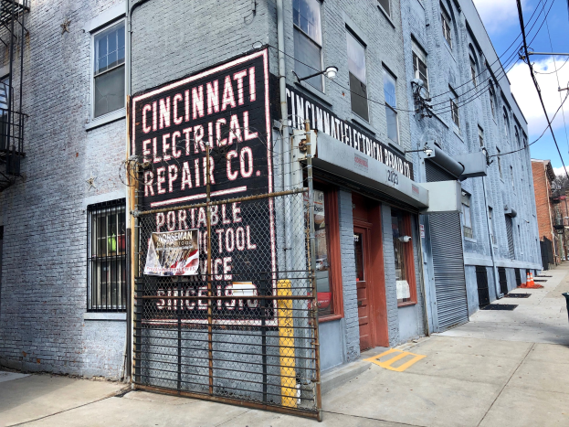 Außenansicht des Cincinnati Electrical Repair Co.-Gebäudes mit einem Schild, einem Metallzaun, einem Müllbehälter, Strommasten, Kabeln, Bäumen und einem bewölkten Himmel.