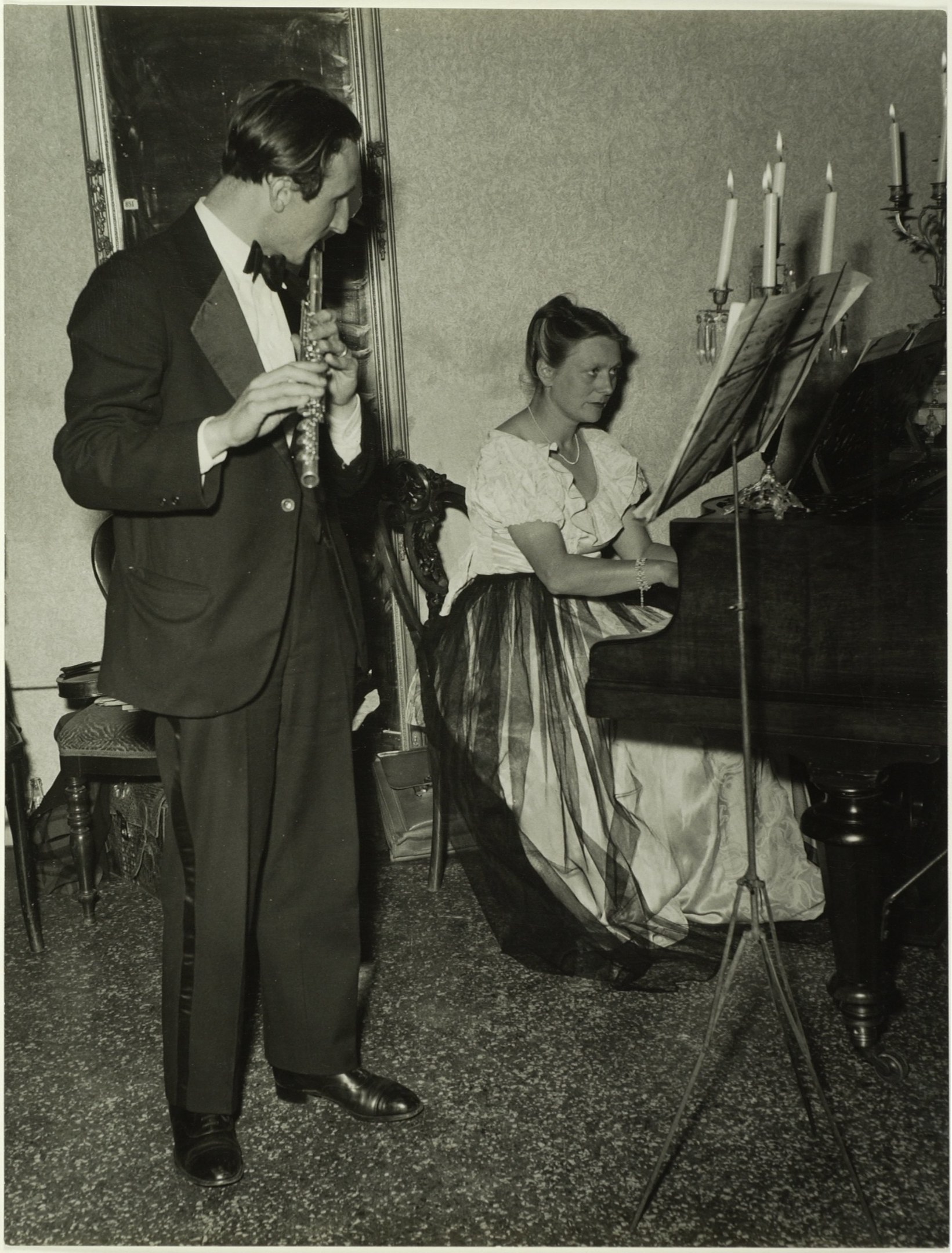 Ein Mann im Smoking spielt Klarinette, während er neben einer Frau steht, die an einem Piano sitzt und Noten auf dem Ständer hat, und ein gerahmtes Bild hängt an der Wand dahinter.