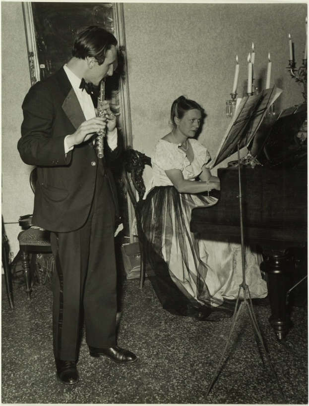 Ein Mann im Smoking spielt Klarinette, während er neben einer Frau steht, die an einem Piano sitzt und Noten auf dem Ständer hat, und ein gerahmtes Bild hängt an der Wand dahinter.