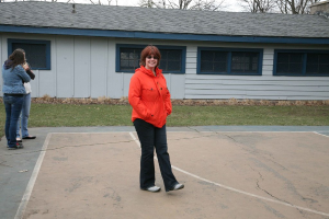 Zwei Personen auf einem Court, eine Frau in einem orangen Shirt in der Mitte, Gras neben dem Court, ein Gebäude mit Dach und Fenstern, Bäume und Himmel im Hintergrund.