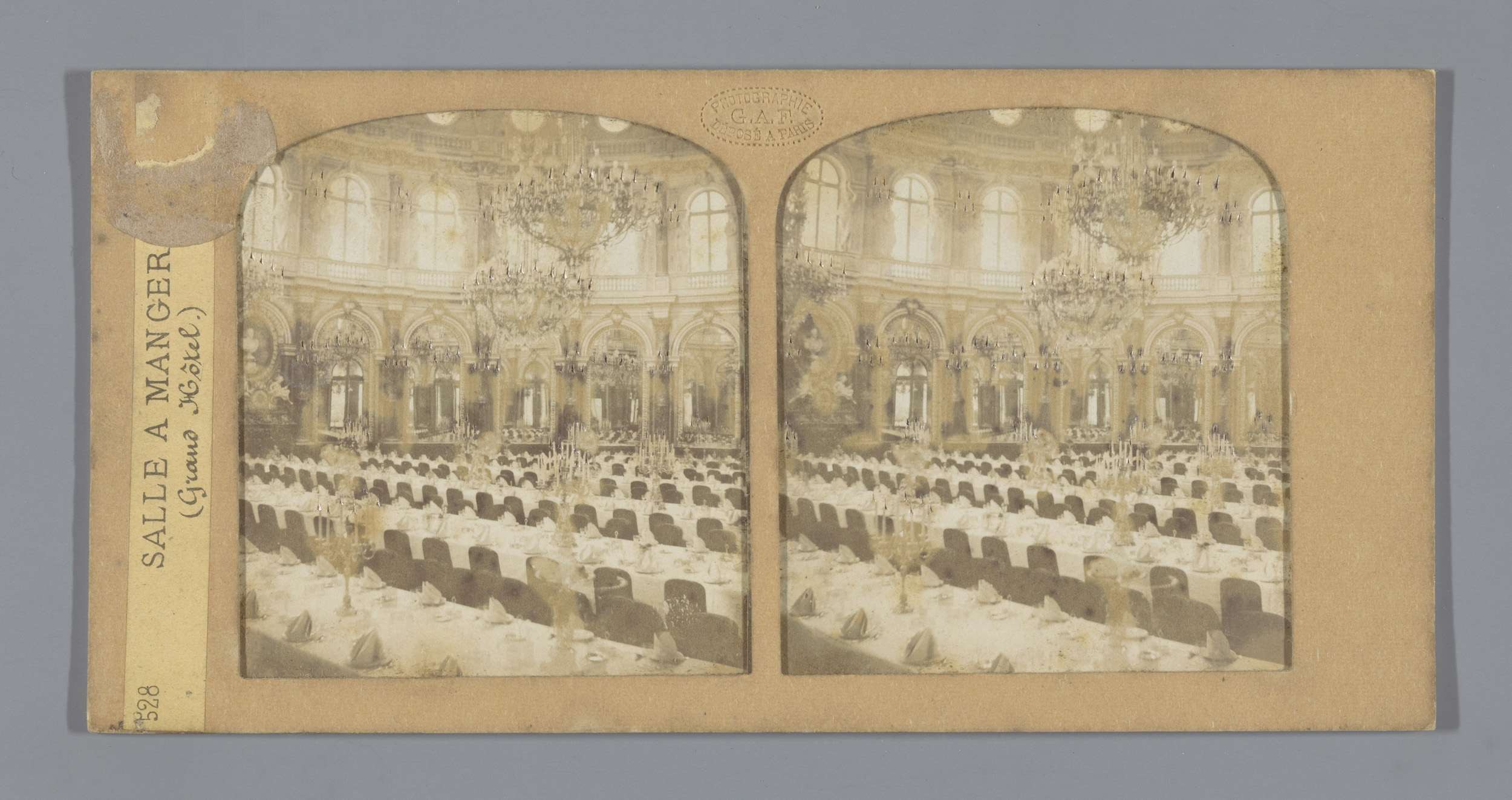 Ein altes Foto eines prächtigen Hotel-Speisesaals mit Tischen, Stühlen, Kronleuchtern und Fenstern, das den Text 'Sale a Manger, Grand Hotel' auf der linken Seite zeigt.
